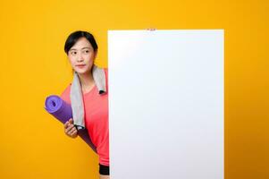 young asian sports fitness woman happy smile wearing pink sportswear standing behind the white blank banner or empty space advertisement board against yellow background. wellbeing lifestyle concept. photo