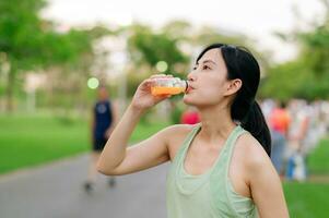 Female jogger. Fit young Asian woman with green sportswear drinking organic orange juice after running and enjoying a healthy outdoor. Fitness runner girl in public park. Wellness being concept photo