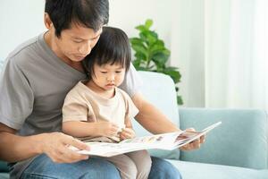 contento asiático padre relajarse y leer libro con bebé hora juntos a hogar. padre sentar en sofá con hija y leyendo un historia. aprender desarrollo, cuidado de niños, reír, educación, contar historias, práctica. foto