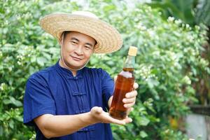 Asian man farmer wears hat, blue shirt and holds bottle of honey at his garden. Concept, Organic agriculture product from bee raising farm or nature sources. Thai local lifestyle. Natural beverage. photo