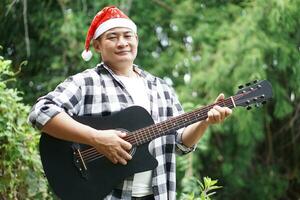 Asian man is playing acoustic guitar in the park, outdoor nature background. Concept, love music, hobby, recreation activity. Relax time. Playing music for celebrate Christmas party. photo