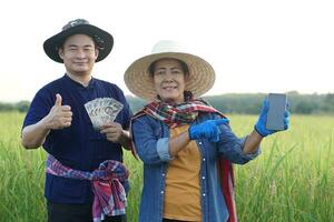 Happy Asian farmers are at paddy field, the man hold Thai banknote money, the woman hold smartphone. Concept, Agriculture occupation. Thai farmers. Profit, income. Online apps for promote products photo