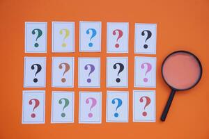 Paper cards of colorful question marks and magnifying glass on orange background. Concept. Teaching aid. Education materials for doing activity or playing investigation games about finding answers. photo