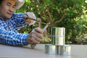 Asian man holds slingshot to play shooting game, aim to shoot the cans in forest. Concept, recreation activity, Hand-powered projectile weapon. Check the accuracy of target shooting. Fun game. photo