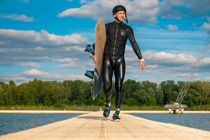 Confident wakeboarder walking with board along pier after training photo