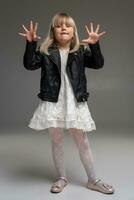 Little blonde girl in a white dress and black leather jacket is posing standing over a gray background. photo