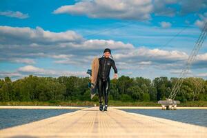Athletic man in wetsuit carrying wakeboard, walking on pier after training photo
