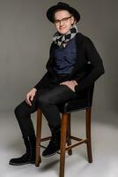 Brunet man in a casual dark clothes posing over a grey background sitting on the bar chair. photo