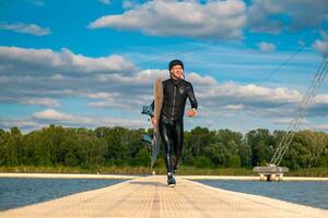 Confident man in wetsuit with wakeboard walking on floating bridge after training photo