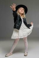 Little blonde girl in a white dress and black leather jacket is posing standing over a gray background. photo