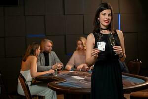 Smiling brunette with glass of champagne and cards in hands standing near poker table photo