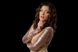 Brunette woman with a curly hair, wearing in a sexy shiny golden dress. She is posing standing against a black studio background. Close-up shot. photo