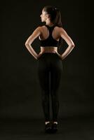 Brunette woman in black leggings, top and sneakers is posing against a black background. Fitness, gym, healthy lifestyle concept. Full length. photo