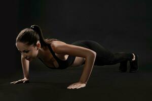 Brunette woman in black leggings, top and sneakers is posing against a black background. Fitness, gym, healthy lifestyle concept. Full length. photo