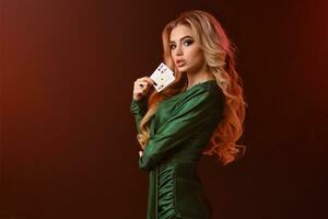 Blonde model in green dress and jewelry. Looking at you, showing two playing cards, posing sideways on brown background. Poker, casino. Close-up photo