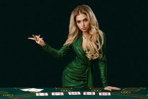 Girl in stylish dress is showing something, leaning on playing table with cards on it, posing on green background. Poker, casino. Close-up, copy space photo