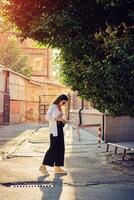 Portrait of a girl in dark sunglasses posing in city. Dressed in top with floral print, white shirt, black trousers, waist bag and sneakers. photo