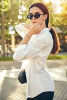 Portrait of a girl in dark sunglasses posing in city. Dressed in top with floral print, white shirt, black trousers, waist bag. photo