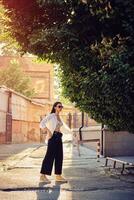 Portrait of a girl in dark sunglasses posing in city. Dressed in top with floral print, white shirt, black trousers, waist bag and sneakers. photo