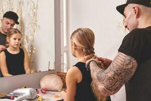 linda pequeño hija y su tatuado papá son jugando juntos cerca un espejo. papá es haciendo su hija cabello. familia fiesta y unión foto