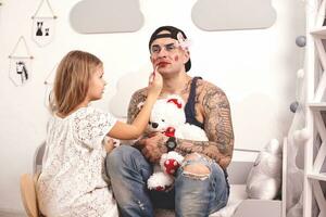 Funny time Tattoed father in a cap and his child are playing at home. Cute girl is doing makeup to her dad in her bedroom. Family holiday togetherness photo