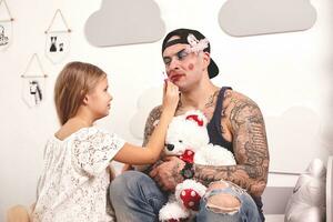 Funny time Tattoed father in a cap and his child are playing at home. Cute girl is doing makeup to her dad in her bedroom. Family holiday togetherness photo