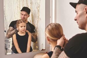 Cute little daughter and her tattoed dad are playing together near a mirror. Dad is doing his daughter's hair. Family holiday and togetherness. photo