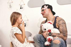 Funny time Tattoed father in a cap and his child are playing at home. Cute girl is doing makeup to her dad in her bedroom. Family holiday togetherness photo