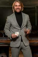 alegre hombre con par de ases y vaso de bebida en pie cerca póker mesa en casino foto