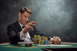Handsome emotional man is playing poker sitting at the table in casino against a white spotlight. photo