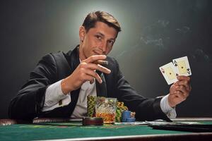 Handsome emotional man is playing poker sitting at the table in casino against a white spotlight. photo