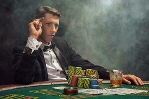 Handsome emotional man is playing poker sitting at the table in casino against a white spotlight. photo