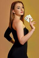 Blonde female with bright make-up, in black dress is showing two aces, posing sideways against colorful background. Gambling, poker, casino. Close-up. photo