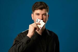 Newbie in poker, in black vest and shirt. Holding two playing cards while posing against blue studio background. Gambling, casino. Close-up. photo