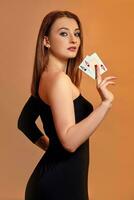 Blonde girl with bright make-up, in black dress is showing two aces, posing sideways against colorful background. Gambling, poker, casino. Close-up. photo