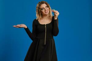 Blonde female in black dress is smiling, showing some colorful chips, posing against blue background. Gambling entertainment, poker, casino. Close-up. photo