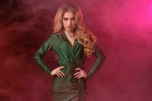 Blonde curly woman in green stylish dress and jewelry. She has put her hands on waist, posing on colorful smoky background. Fashion, beauty. Close up photo