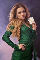 Blonde model in green dress and jewelry. Put hand on hip, showing two playing cards, posing on purple smoky background. Poker, casino. Close-up photo