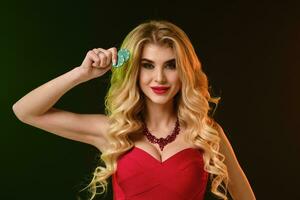 Blonde lady in red fitting dress and necklace. She smiling, showing two green chips, posing on colorful background. Poker, casino. Close-up photo