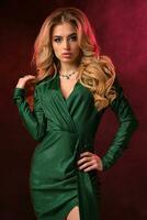 Blonde curly female in green stylish dress and jewelry. She has put her hand on waist, posing on colorful smoky background. Fashion, beauty. Close up photo