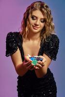 Blonde girl in black sequin dress is showing handful of multicolored chips, posing on colorful studio background. Gambling, poker, casino. Close-up. photo