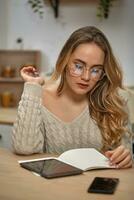 Girl blogger in glasses, beige sweater. Sitting in kitchen at table with smartphone and tablet on it. Going to write something in notebook. Close-up photo