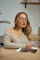 Female blogger in glasses, beige sweater. Sitting in kitchen at wooden table with cup and tablet on it. Going to write something in notebook. Close-up photo