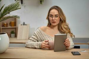 Female blogger in glasses, beige sweater. Holding tablet, pink cup. Sitting in kitchen at wooden table with palm, smartphone, plastic card. Close-up photo