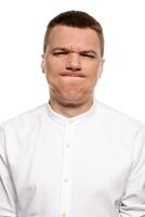 Charming handsome young man in a white shirt is making faces, while standing isolated on a white background photo