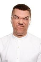 Charming handsome young man in a white shirt is making faces, while standing isolated on a white background photo