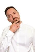Portrait of unhappy determined european male with bristle touching chin while thinking and looking with serious and worried look at camera, standing against white background. photo