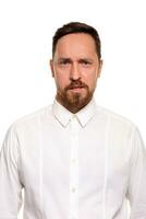 Portrait of handsome man with stubble has indignant expression, frowns face, dressed in white shirt, isolated over white background. photo