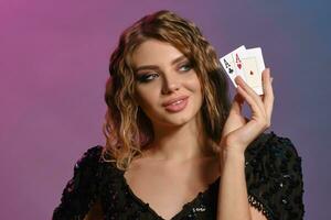 Brown-haired woman in black shiny dress is smiling, showing two playing cards, posing on colorful background. Gambling, poker, casino. Close-up. photo