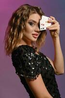 Brown-haired girl in black shiny dress showing two aces, posing sideways on colorful background. Gambling entertainment, poker, casino. Close-up. photo